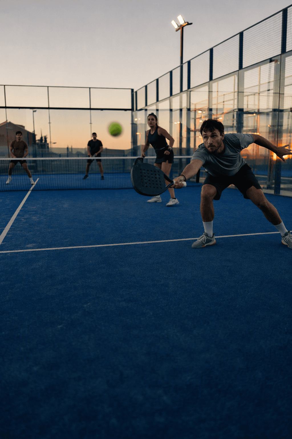 Four friends mid-rally on a glass padel court at dusk